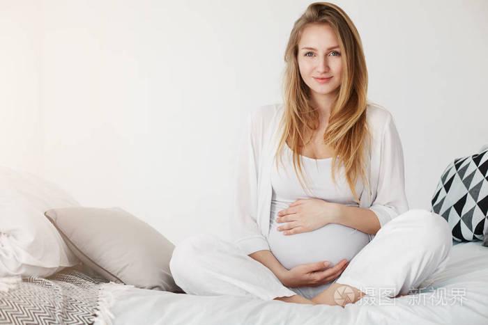 健康怀孕妊娠一名年轻孕妇微笑着坐在她的卧室休息早上抱着她的肚子